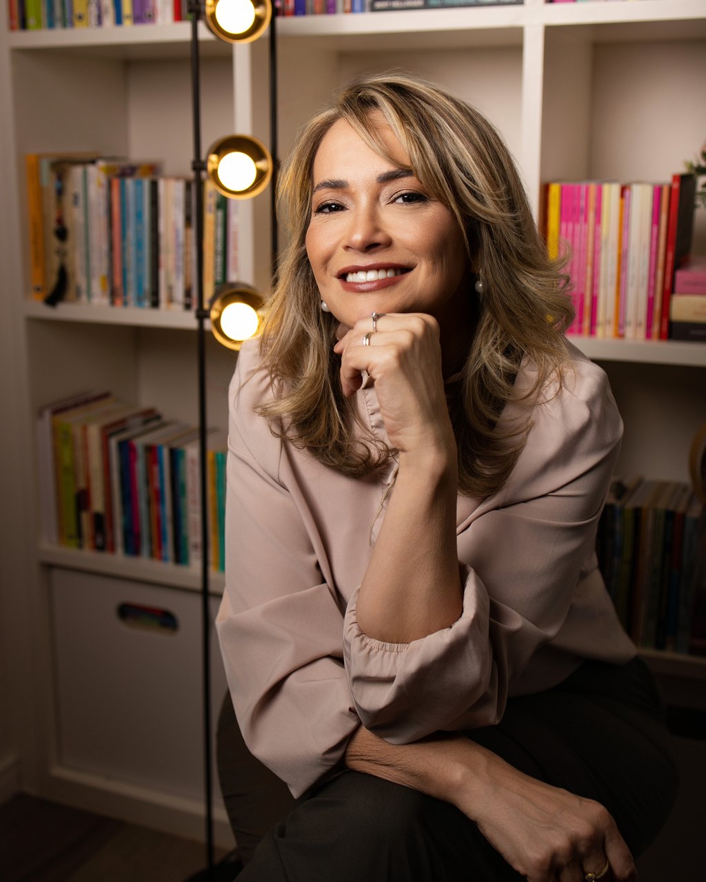 A psicóloga Cinthia Sàntiago sorrindo, com a mão apoiada no queixo, em frente a uma estante de livros no consultório dela.
